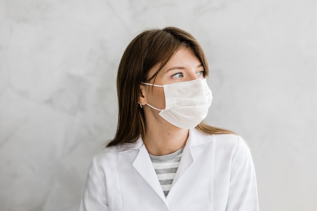 Primo piano di una dottoressa caucasica con maschera facciale in un abito medico bianco su sfondo grigio. Ritratto di dottoressa o infermiere in ospedale. Personale sanitario, salute, concetto di medicina