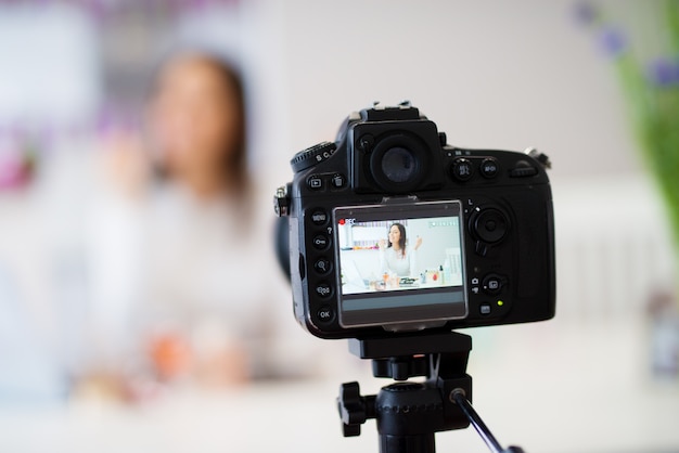 Primo piano di una fotocamera per scattare foto della giovane bella ragazza carina che è seduto al tavolo della cucina con un computer portatile su di esso e mostrando profumi e cosmetici alla fotocamera mentre sorride.