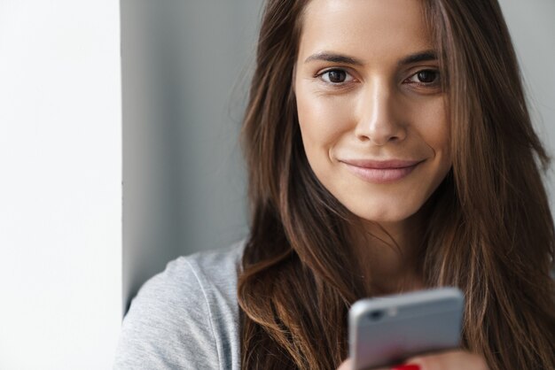 Primo piano di una giovane bella ragazza sorridente in piedi appoggiata a un muro grigio su un muro grigio, usando il telefono cellulare