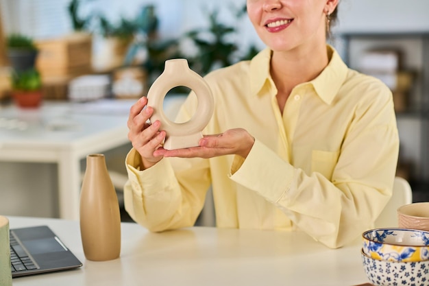 Primo piano di una giovane blogger felice che presenta un nuovo vaso in ceramica fatto a mano di colore beige mentre è seduto davanti alla telecamera