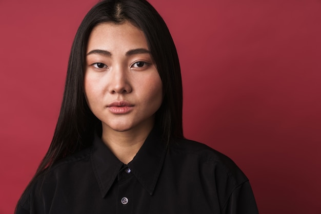 Primo piano di una giovane donna asiatica abbastanza sicura di sé che indossa abiti casual in piedi isolata sul muro rosso, guardando la telecamera