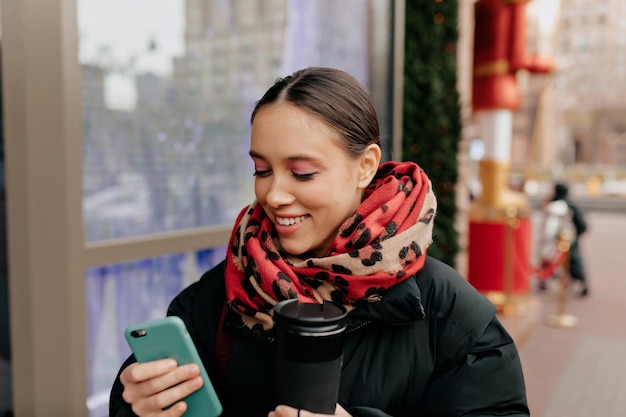 Primo piano di una giovane donna attraente con i denti che usa lo smartphone in mano all'aperto Ragazza alla moda che sorride mentre guarda lo schermo dello smartphone Concetto di persone dipendenti