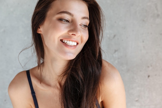 Primo piano di una giovane donna bruna sorridente che indossa un reggiseno isolato su un muro grigio