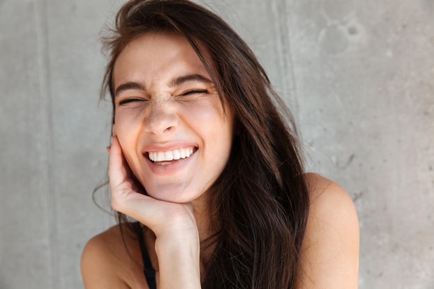 Primo piano di una giovane donna bruna sorridente che indossa un reggiseno isolato sul muro grigio, ridendo