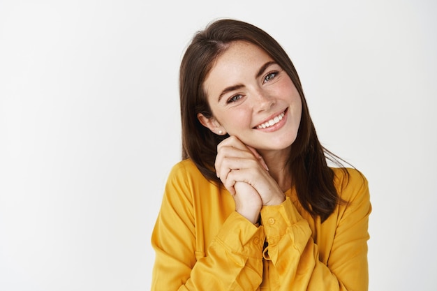 Primo piano di una giovane donna carina che sembra felice alla telecamera, tenendo le mani insieme vicino al viso e sorridendo, ammirando qualcosa, in piedi sul muro bianco