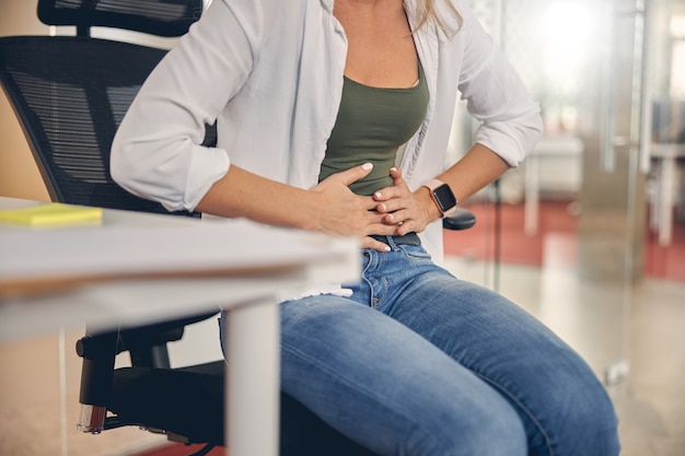 Primo piano di una giovane donna che sente dolore allo stomaco mentre è seduta su una sedia al lavoro