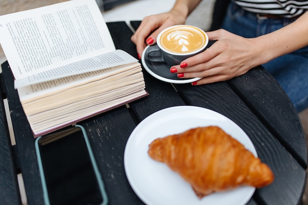 Primo piano di una giovane donna che tiene in mano un caffè con un libro di carta e un croissant su un tavolo da terrazza