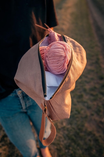 Primo piano di una giovane donna che tiene una borsa fatta a mano con accessori per lavorare a maglia ferri da maglia uncinetti palla rosa di filato sullo sfondo di un campo