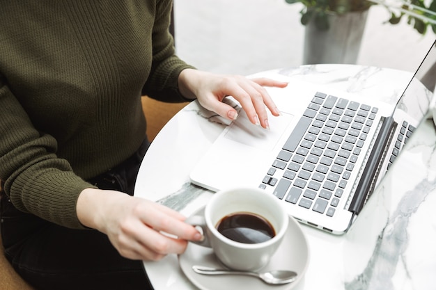 Primo piano di una giovane donna dai capelli rossi che si rilassa al tavolino del bar all'interno, beve caffè, lavora al computer portatile