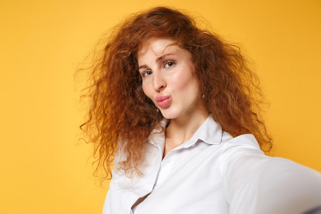 Primo piano di una giovane donna dai capelli rossi in camicia bianca in posa isolata sul muro giallo arancione