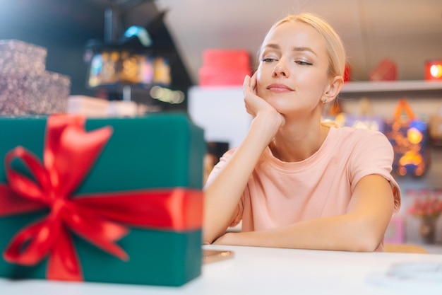 Primo piano di una giovane donna infelice seduta di fronte a una scatola regalo di natale che lega un nastro rosso e