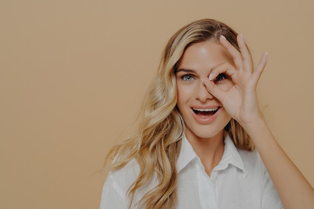 Primo piano di una giovane donna positiva con lunghi capelli biondi ondulati che guarda l'obbiettivo attraverso le dita piegate in un gesto ok, apre la bocca con eccitazione mentre posa isolata su sfondo beige