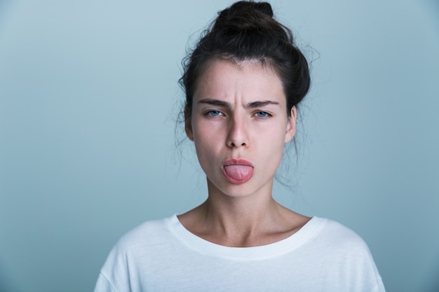 Primo piano di una giovane donna sconvolta che indossa t-shirt isolate su sfondo blu, con la lingua fuori