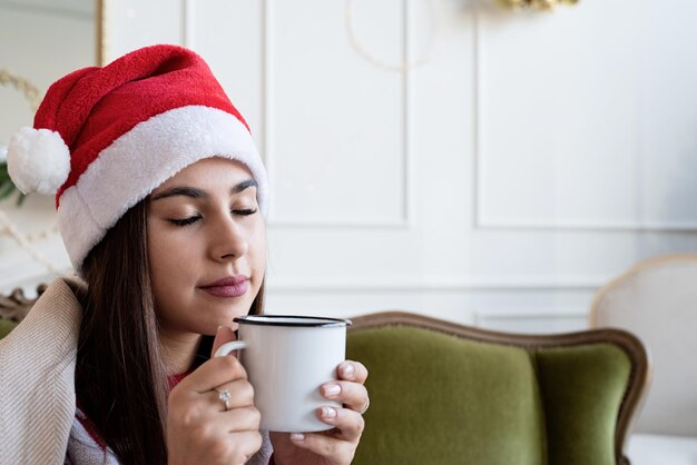 Primo piano di una giovane donna seduta sul divano da sola in un soggiorno decorato per il Natale con una tazza di bevanda calda