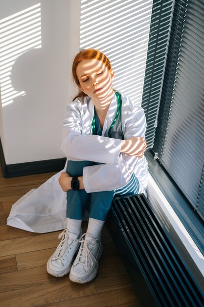 Primo piano di una giovane dottoressa triste frustrata in camice bianco seduta sul pavimento che abbraccia le gambe con le mani vicino alla finestra.