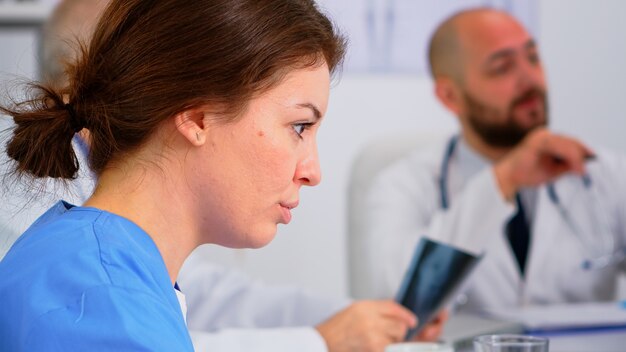 Primo piano di una giovane infermiera che discute con un collega durante una conferenza medica seduta sulla scrivania nell'ufficio riunioni dell'ospedale. Parlare dei sintomi della malattia nella stanza della clinica durante il brainstorming