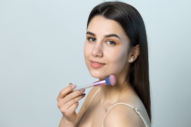 Primo piano di una giovane ragazza in un top leggero su uno sfondo bianco che fa un trucco facciale. Una bella donna tiene un pennello cosmetico vicino al viso e sorride.