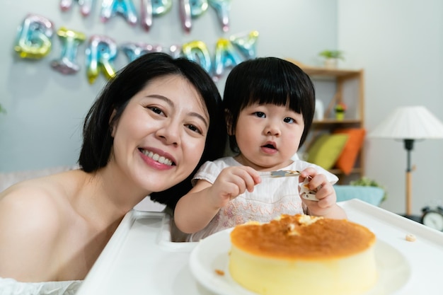 primo piano di una madre coreana abbastanza felice che sorride alla telecamera con la testa vicino alla figlia del bambino che sta mangiando la torta alla festa di compleanno a casa.