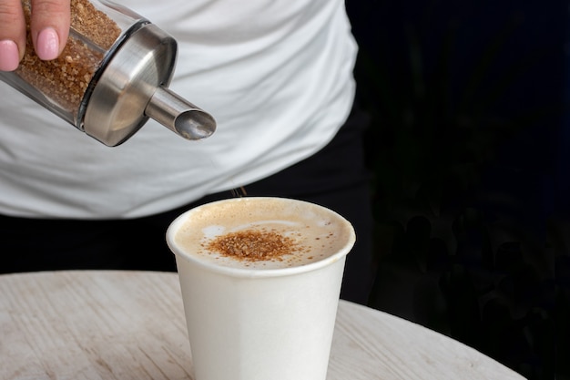 Primo piano di una mano che versa lo zucchero di canna in una tazza di plastica bianca con caffè