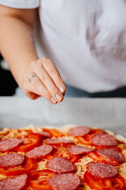 Primo piano di una mano che versa pepe ed erbe aromatiche in una pizza ai peperoni. Preparare cibo fatto in casa per incontrare gli amici.