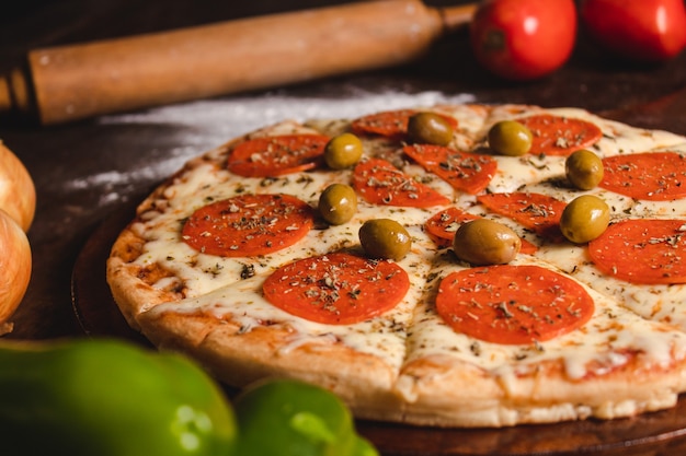 Primo piano di una pizza ai peperoni con olive e diverse verdure intorno sopra un tavolo di legno.