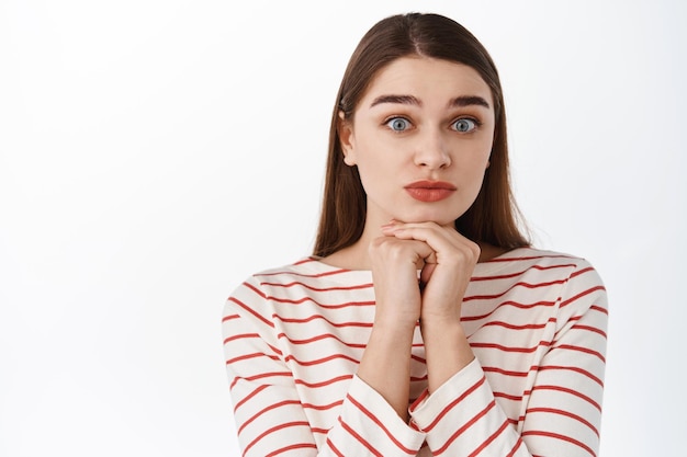 Primo piano di una ragazza carina e timida, tenere le mani vicino al viso, desiderare e implorare qualcosa, fare una faccia stupida per chiedere un favore, in piedi su sfondo bianco