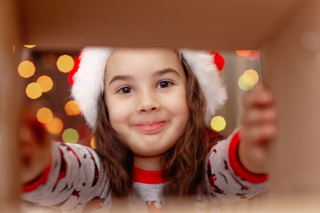 Primo piano di una ragazza felice in un cappello di Babbo Natale prende un regalo da una scatola, vista interna