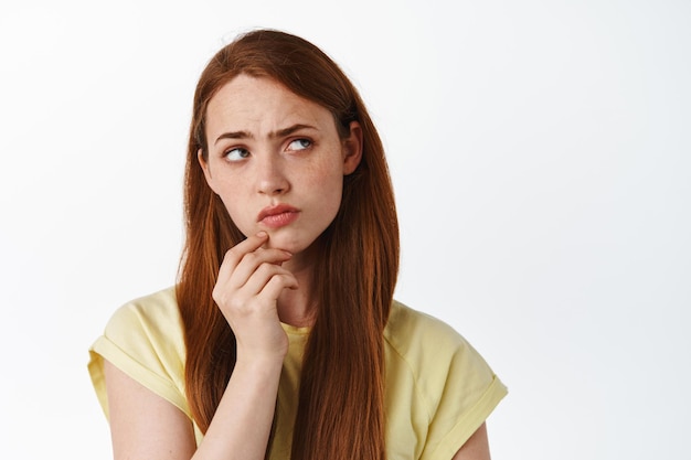 Primo piano di una ragazza rossa seria premurosa, toccare il mento e guardare da parte, pensare a qualcosa, avere ipotesi, essere sospettoso, sfondo bianco.