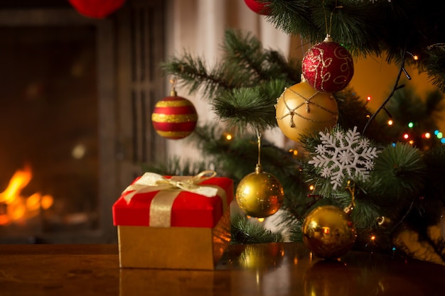 Primo piano di una scatola regalo di Natale rossa con nastro dorato accanto all'albero di Natale decorato e al caminetto acceso