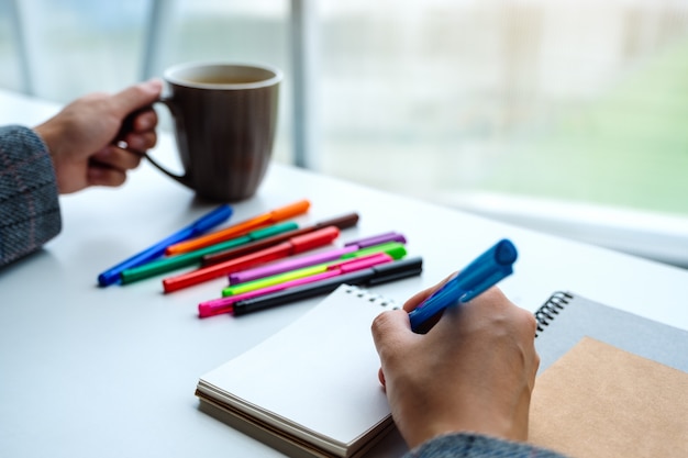 Primo piano di una scrittura della donna su un taccuino in bianco con le penne colorate mentre bevendo caffè