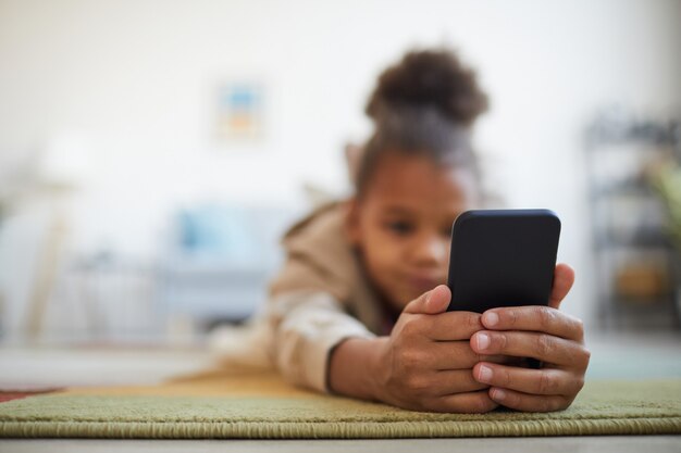Primo piano di una simpatica ragazza afro-americana che tiene in mano lo smartphone mentre è sdraiato sul pavimento in un accogliente interno di casa, copia spazio