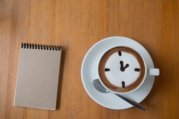 Primo piano di una tazza calda caffè latte arte disegnato un concetto di business oclock sul tavolo di legno con il libro di nota