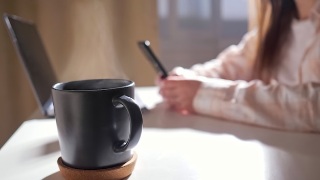 Primo piano di una tazza con bevanda calda sul tavolo sullo sfondo di una donna irriconoscibile con telefono e laptop zoom out