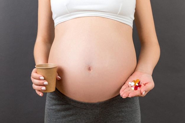 Primo piano di una tazza di carta e di un mucchio di pillole nelle mani di una donna incinta su sfondo colorato con spazio per la copia Concetto di assistenza sanitaria