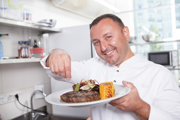 Primo piano di uno chef maschio felice sorridente che sala la bistecca di manzo su una piastra