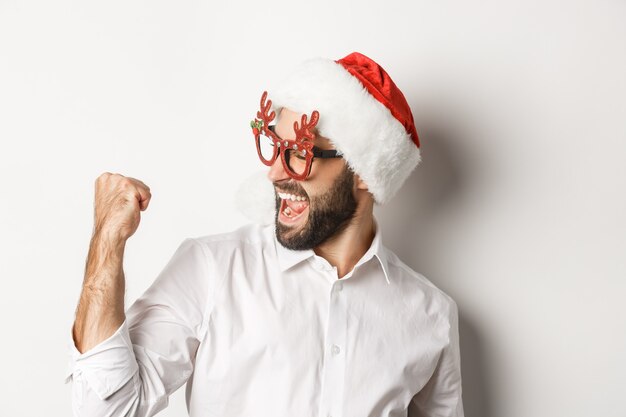 Primo piano di uomo barbuto felice che indossa cappello da Babbo Natale e occhiali da festa di Natale, facendo pompare il pugno e gioire, raggiungere l'obiettivo e celebrare, sfondo bianco.