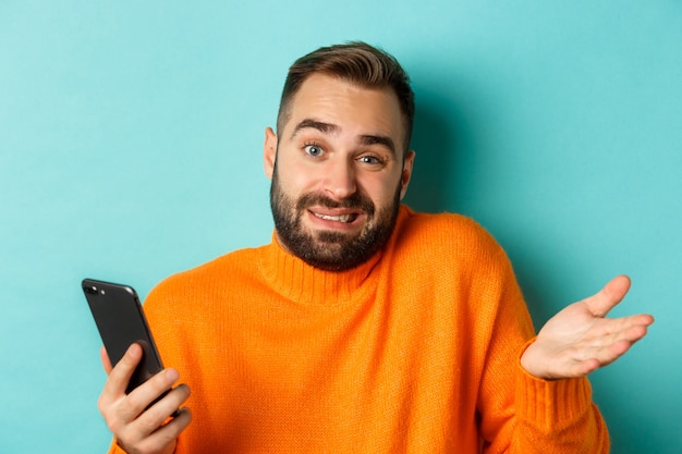 Primo piano di uomo confuso che scrolla le spalle durante l'utilizzo del telefono, in piedi indeciso su sfondo turchese.