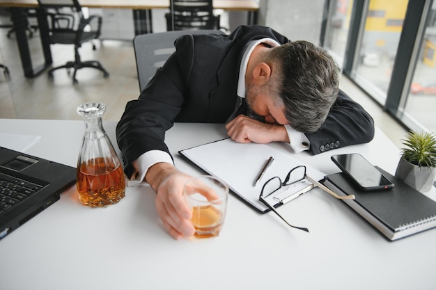 Primo piano di uomo d'affari stressato con in mano un bicchiere di whisky che dorme e Data Chartsbusiness documento alla scrivania dell'ufficio dipendenza da alcol e concetto di uomo d'affari ubriaco