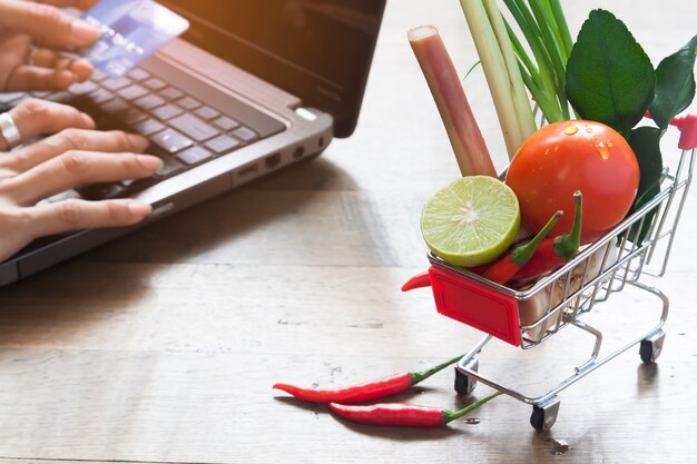 Primo piano di verdure fresche nel carrello della spesa con la donna utilizzando il computer portatile e la carta di credito, shopping online