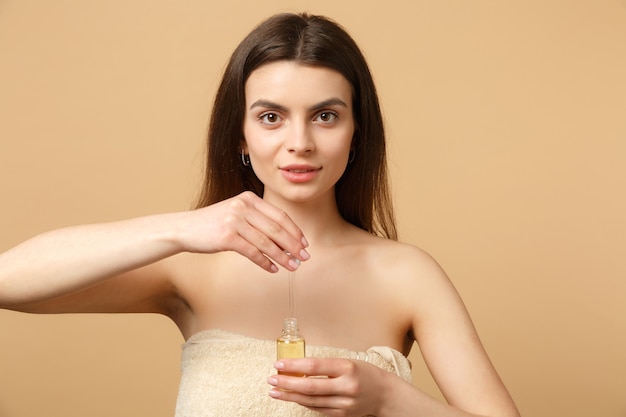 Primo piano donna mezza nuda bruna con pelle perfetta, trucco nudo applicare olio dalla bottiglia sul viso isolato su muro beige pastello