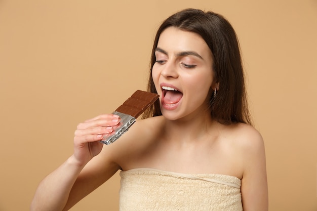 Primo piano donna mezza nuda con pelle perfetta, trucco nudo tiene una barretta di cioccolato isolata su muro beige pastello