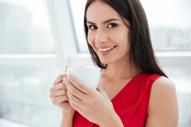 Primo piano donna sorridente con una tazza di caffè in piedi vicino alla finestra in ufficio e guardando la telecamera