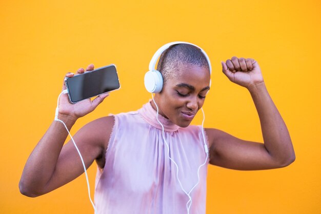 Primo piano e ritratto di bella donna afroamericana che sorride e si diverte a ballare ascoltando musica con le cuffie dal suo telefono - godendosi lo stile di vita della tecnologia