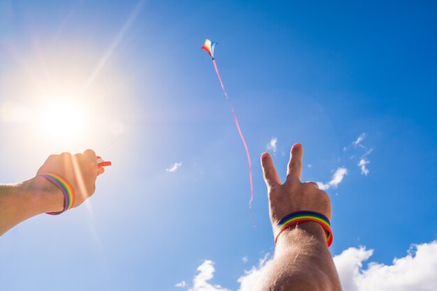 Primo piano e ritratto di braccia e mani in alto che indossano un braccialetto di colori lgtb e fanno volare un aquilone nel cielo