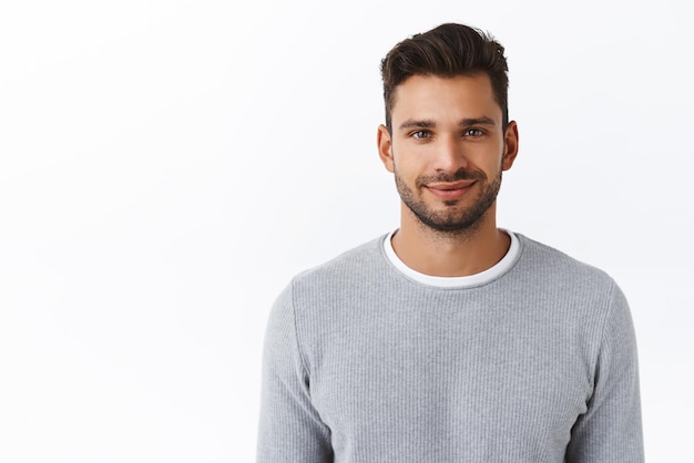 Primo piano felice sorridente fiducioso uomo macho con setole in elegante maglione grigio che sembra affidabile e sincero con un sorriso gentile amichevole in piedi sfondo bianco sicuro di sé e motivato