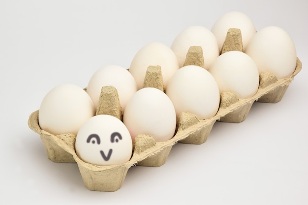Primo piano fotografato di un vassoio laterale con dieci uova di gallina bianca. L'uovo più vicino ha un disegno animato di una faccia sorridente