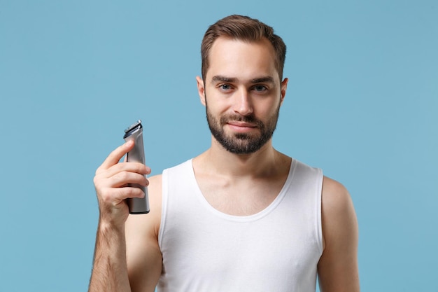 Primo piano giovane barbuto di 20 anni in camicia bianca tenere rasoio elettrico isolato su sfondo blu pastello, ritratto in studio. Concetto di procedure cosmetiche sanitarie per la cura della pelle. Simula lo spazio della copia.