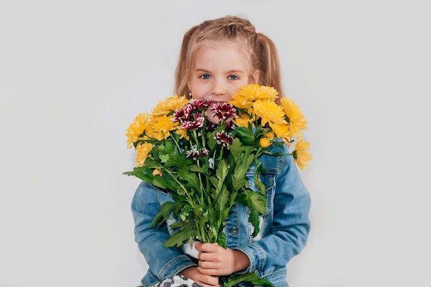 Primo piano, messa a fuoco selettiva. Bambina bionda in una tiene crisantemi e gerbere nelle sue mani su uno sfondo bianco, una bambina sorride e tiene in mano fiori primaverili