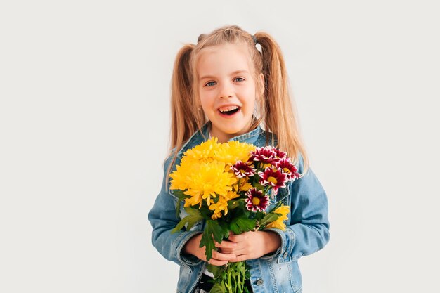 Primo piano, messa a fuoco selettiva. Bambina bionda in una tiene crisantemi e gerbere nelle sue mani su uno sfondo bianco, una bambina sorride e tiene in mano fiori primaverili