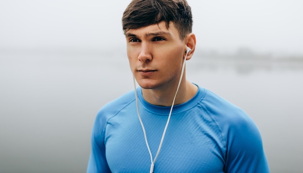 Primo piano orizzontale all'aperto ritratto di giovane corridore in posa durante il jogging al mattino sullo sfondo del lago di nebbia Fitness maschile che si esercita nel parco e ascolta la musica con gli auricolari